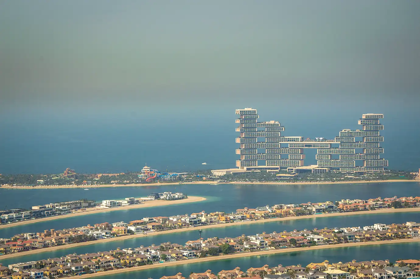 An aerial perspective showing the stacked-block architectural structure of Atlantis The Royal resort on Dubai's Palm Jumeirah crescent, with residential frond villas and blue sea channels in the foreground.