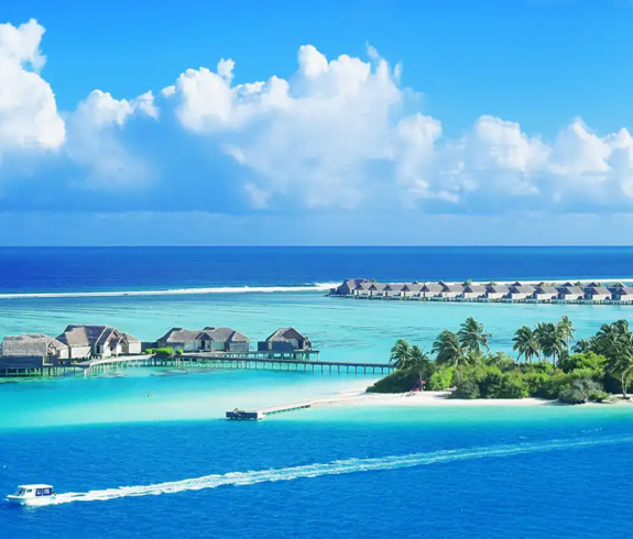 An aerial photograph showing overwater bungalows, a tropical island lined with palm trees, and a white speedboat cruising through deep blue and turquoise ocean waters under a bright sky in the Maldives.