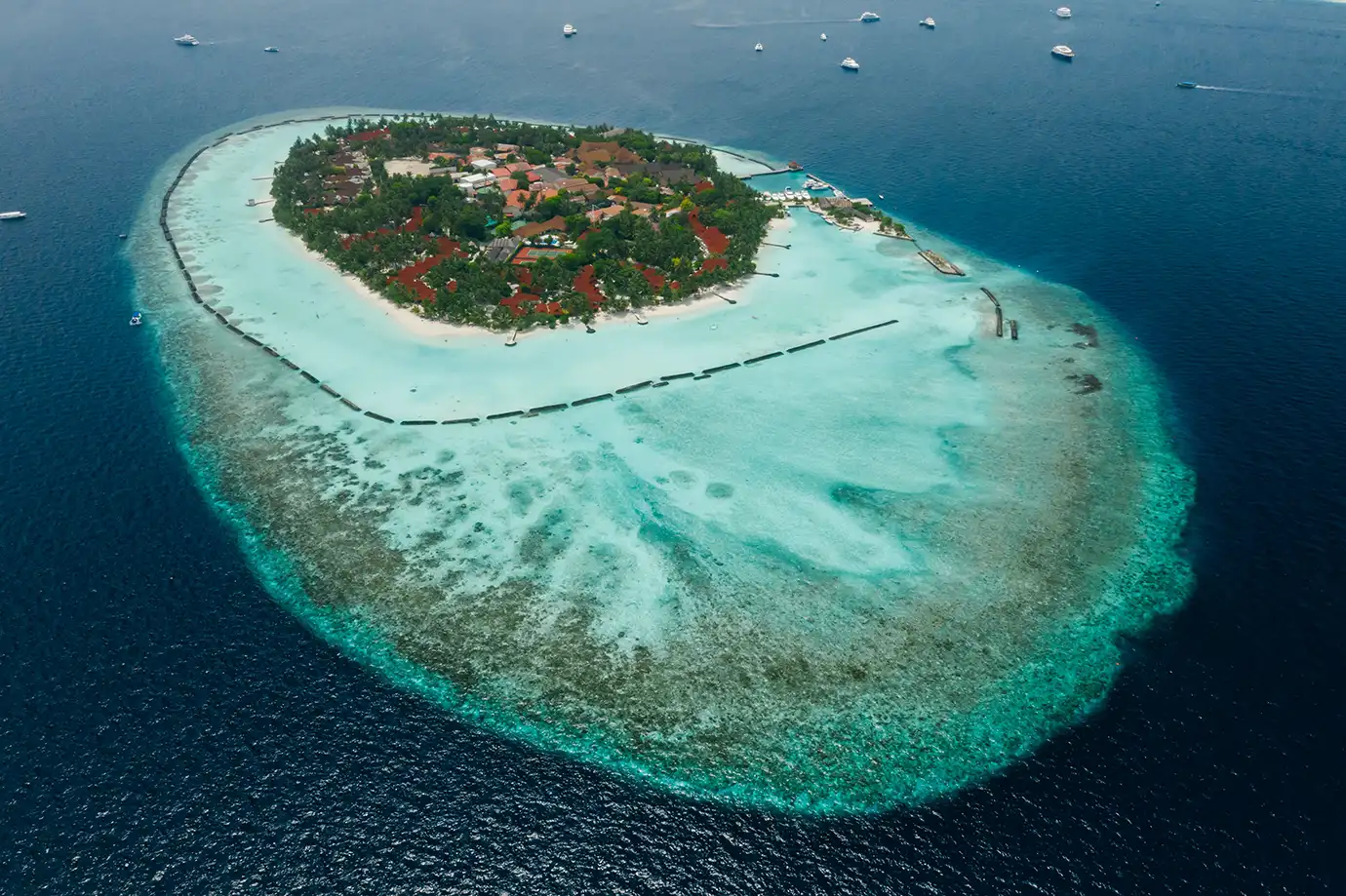 A high-angle aerial photograph looking down on an oval-shaped tropical island resort with white sand beaches, dense green palm foliage, and red roofs, encircled by a shallow turquoise coral lagoon dropping off into the deep blue Indian Ocean in the Maldives. 