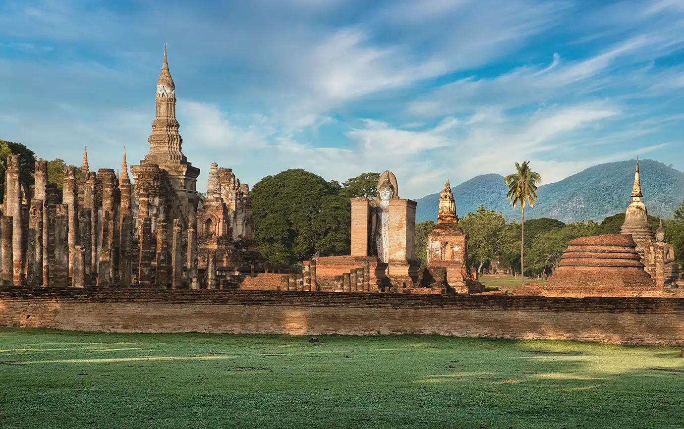 A wide landscape shot of the ancient brick chedi towers, stone pillars, and seated Buddha statues of Wat Mahathat in Sukhothai Historical Park, Thailand, set against distant mountains.