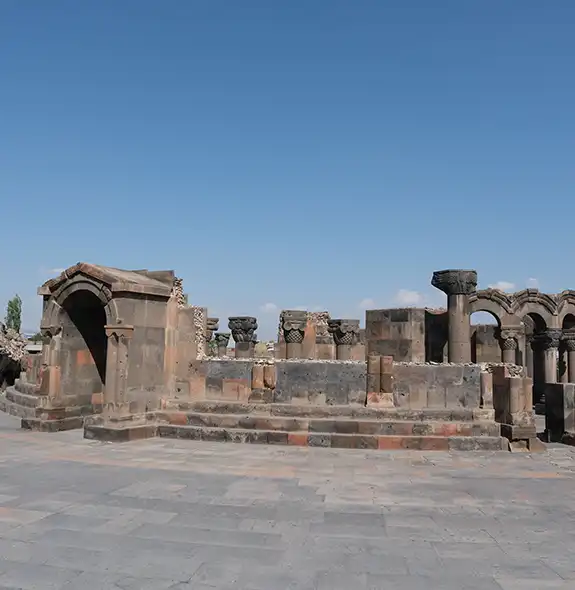 The preserved stone arches, carved pillars, and entrance structure of Zvartnots Cathedral ruins in Armenia, set against a wide paved stone courtyard and a clear blue sky.