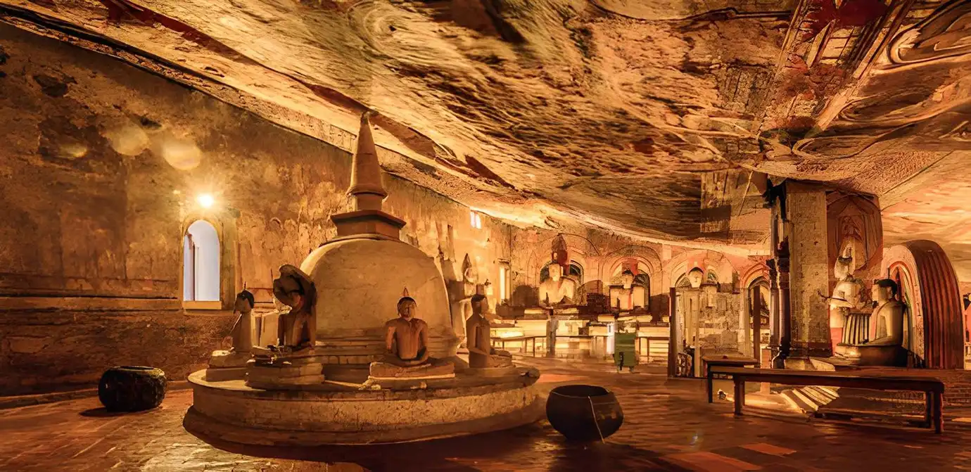 The dimly lit interior of a cave temple in Dambulla, Sri Lanka, featuring a central white stupa surrounded by seated Buddha statues under an arched rock ceiling covered in ancient murals.