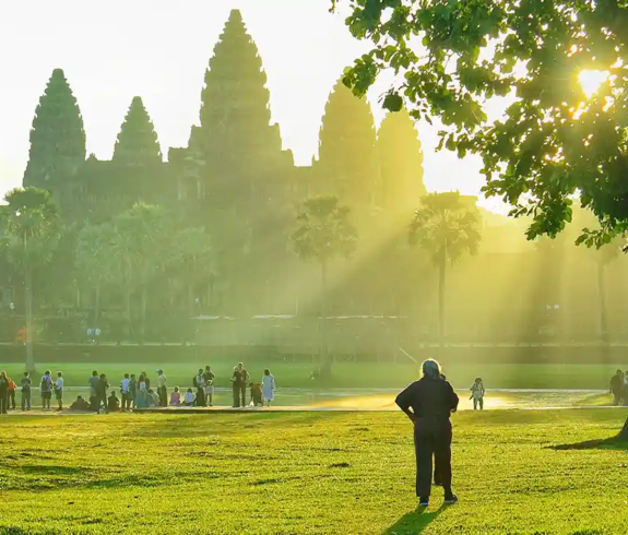 A crowd of international tourists strolling across green lawns near Angkor Wat as sunlight filters through trees featured in a travel guide on Is Cambodia Safe.