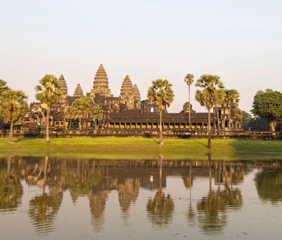 A wide landscape view of the ancient Angkor Wat stone temple complex and surrounding tall sugar palm trees reflecting clearly on the surface of a calm pond under a soft morning sky in Siem Reap, Cambodia.