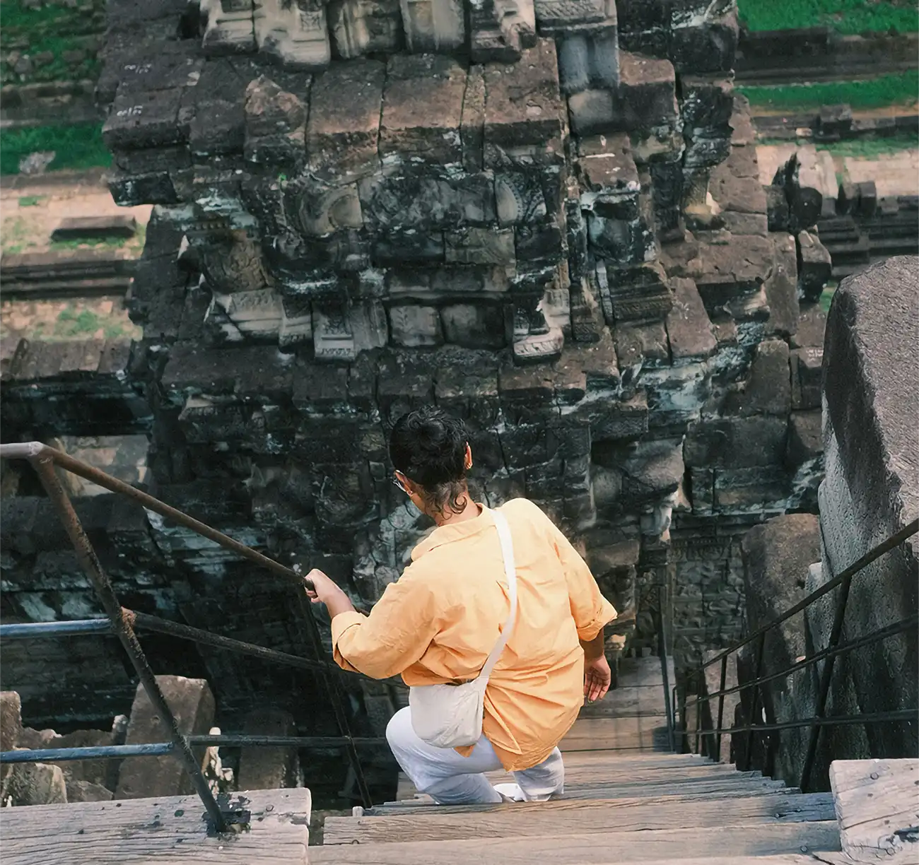 A traveler wearing a respectful long-sleeved shirt and trousers carefully descending steep wooden stairs at an ancient temple tower featured in a travel guide on Angkor Wat Tickets.