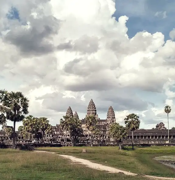 A wide shot of the stone towers of Angkor Wat framed by tall palm trees, green grass fields, and a dirt path under a sky filled with dramatic white and grey clouds in Siem Reap, Cambodia.