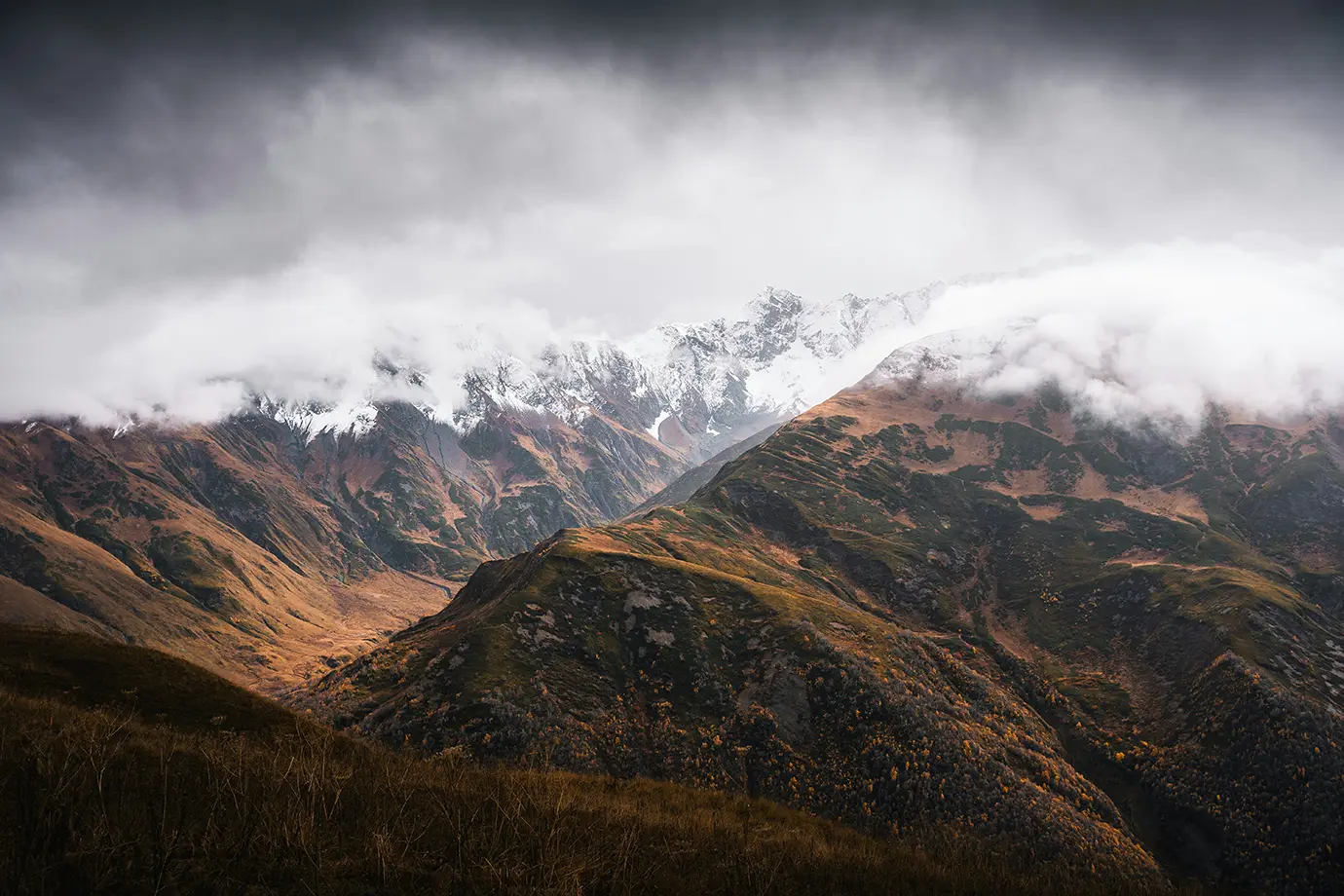 Rolling autumn slopes in golden hues beneath snow-dusted Greater Caucasus mountain peaks shrouded in dramatic clouds, representing the Best Time to Visit Georgia.