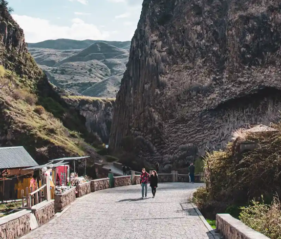 A couple walking down a stone path flanked by towering basalt cliffs leading toward Geghard Monastery featured in a travel guide on When Is the Best Time to Visit Armenia.