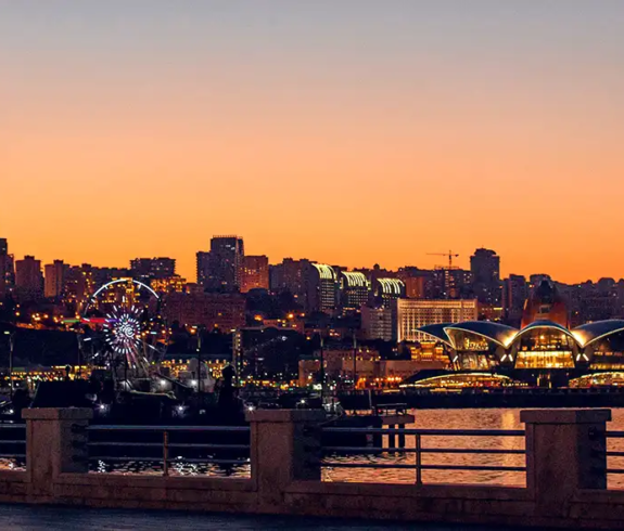 A wide evening view across the paved waterfront promenade looking toward the illuminated Baku city skyline, featuring the red-lit Flame Towers and Ferris wheel against a vibrant orange and dark blue sunset sky in Azerbaijan.