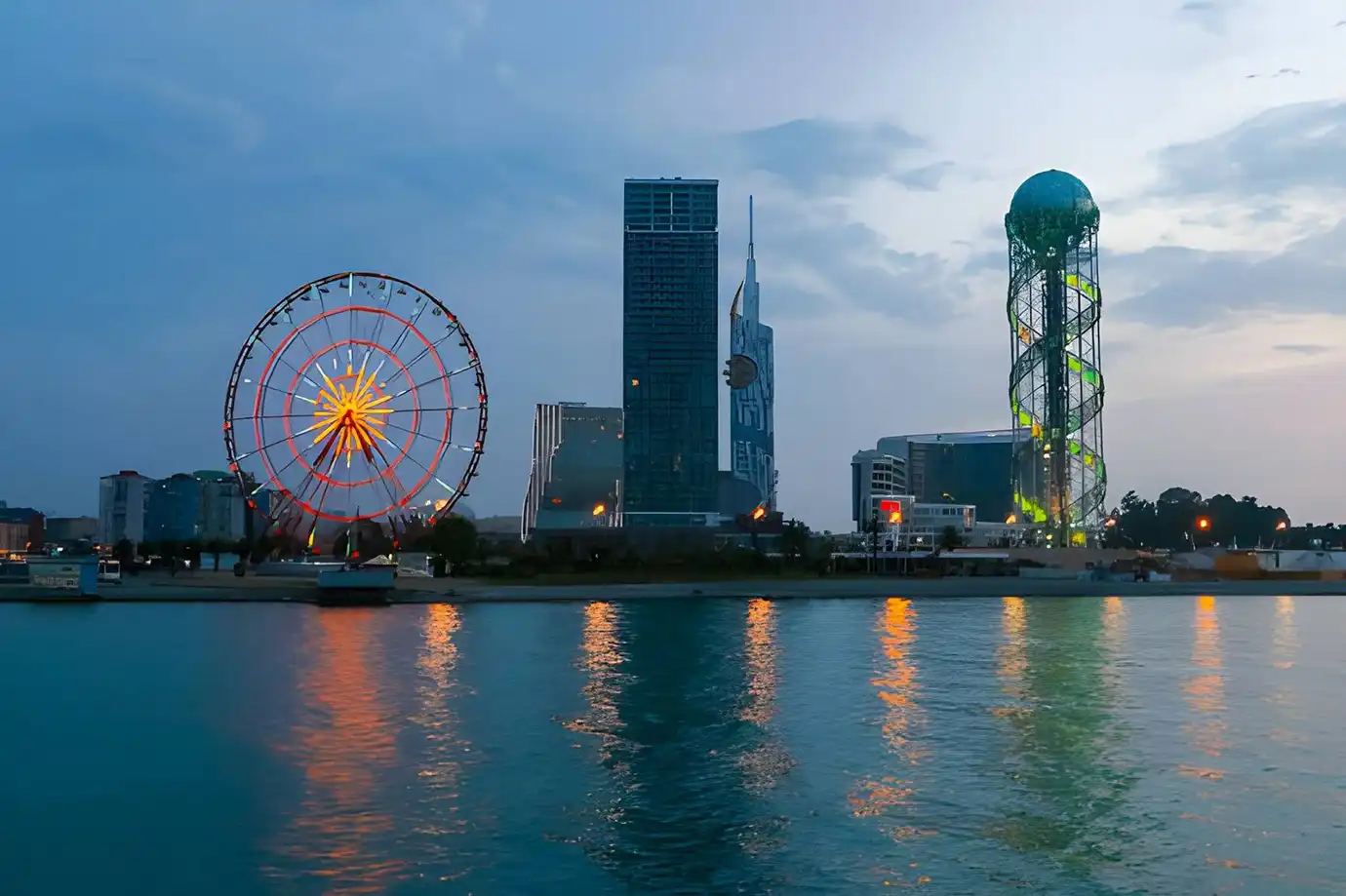 A serene evening view from the water showing the illuminated Batumi waterfront skyline, including the glowing Ferris wheel and Alphabet Tower, for the Best Time to Visit Georgia.