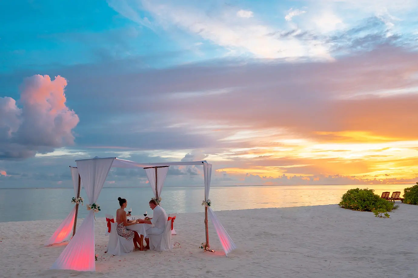 A couple dining under a white canopy on a pristine sandy beach during a golden sunset featured in a guide on How to Plan a Maldives Luxury Honeymoon on an Affordable Budget.
