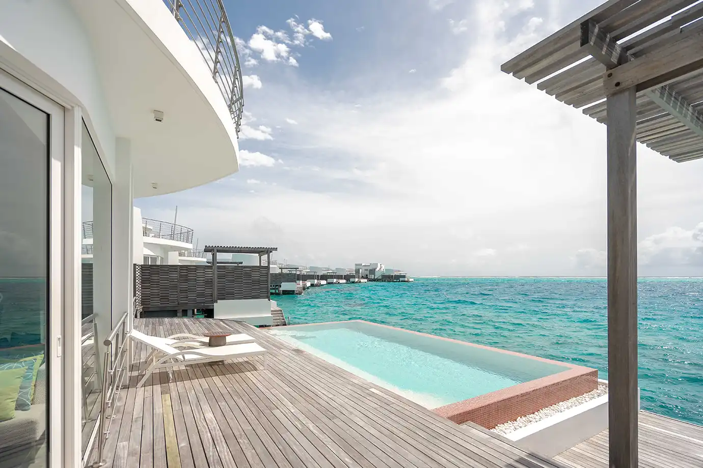 A contemporary overwater resort deck featuring white sun loungers and a private infinity pool overlooking turquoise ocean waters featured in a guide on How to Plan a Maldives Luxury Honeymoon on an Affordable Budget.
