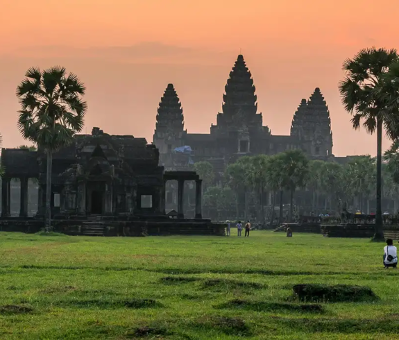 A panoramic view of two travelers sitting on a vibrant green lawn watching the sunrise over Angkor Wat featured in a travel guide on the Best Time to Visit Cambodia.