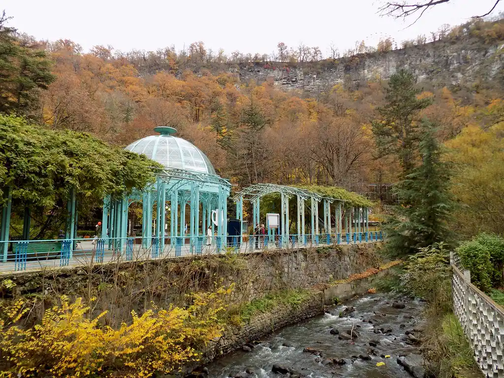A turquoise glass dome pavilion and arcade running alongside a stream in autumn, surrounded by colorful foliage and steep cliffside forest, representing nature-focused Places to Visit in Georgia.