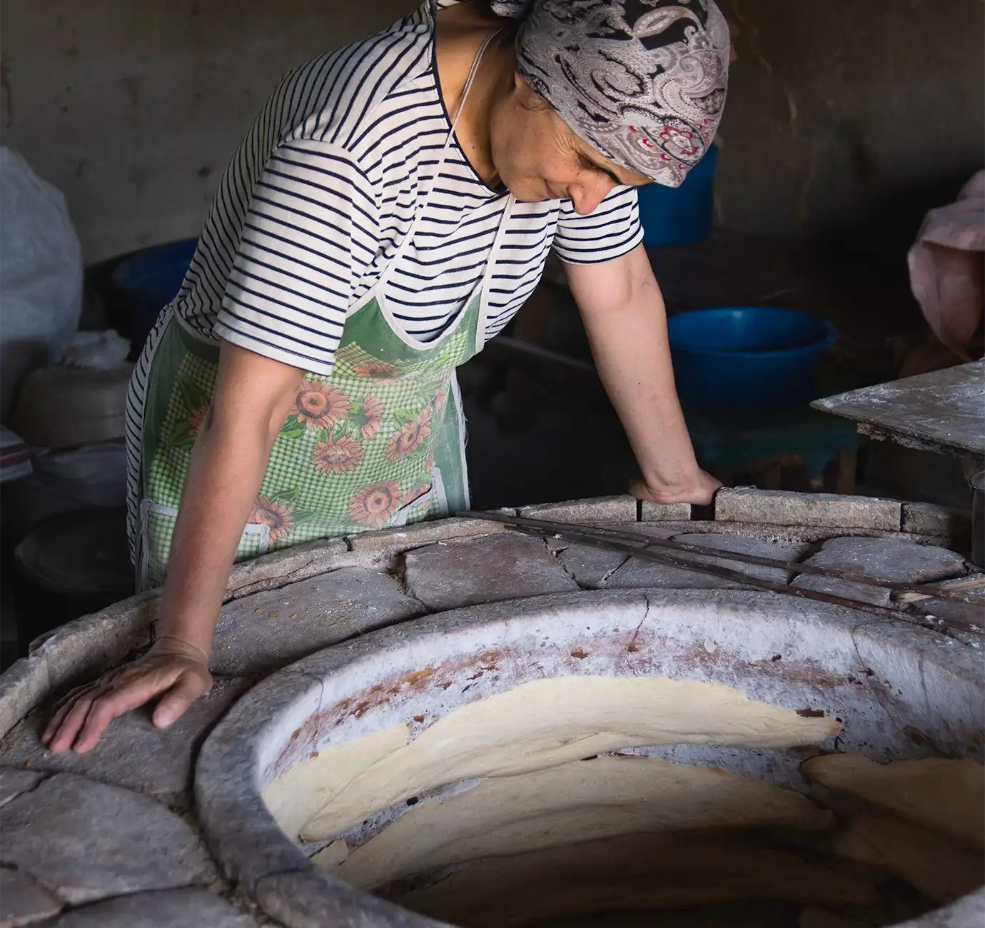 A local woman in a floral apron tending to flatbread baking inside a subterranean clay tonir oven featured in a culinary guide on What to Eat in Armenia.