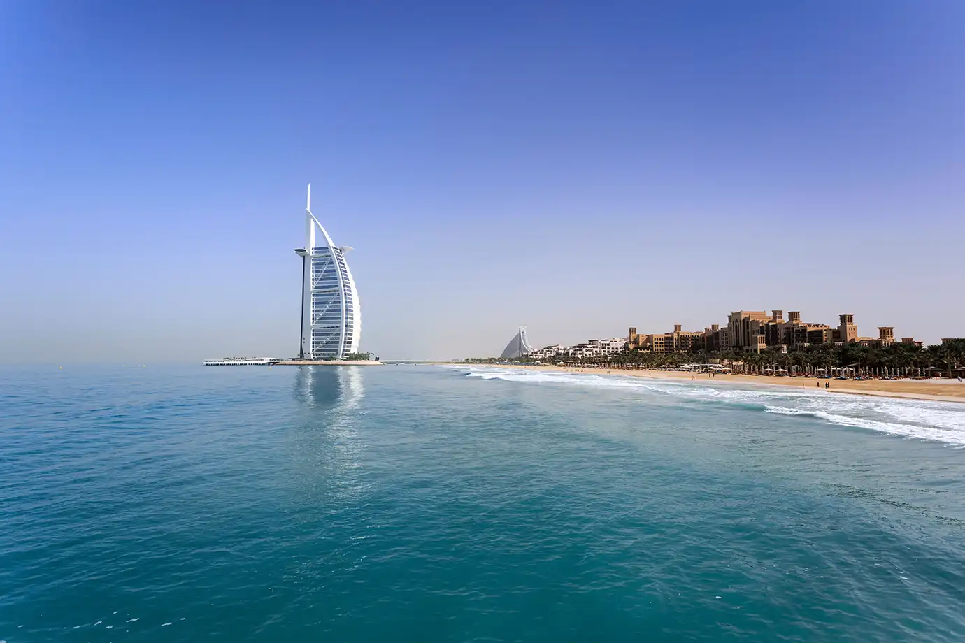 A wide coastal shot showing the sail-shaped Burj Al Arab hotel standing off the shore of a sandy beach resort, surrounded by clear turquoise ocean water under a bright blue sky in Dubai.