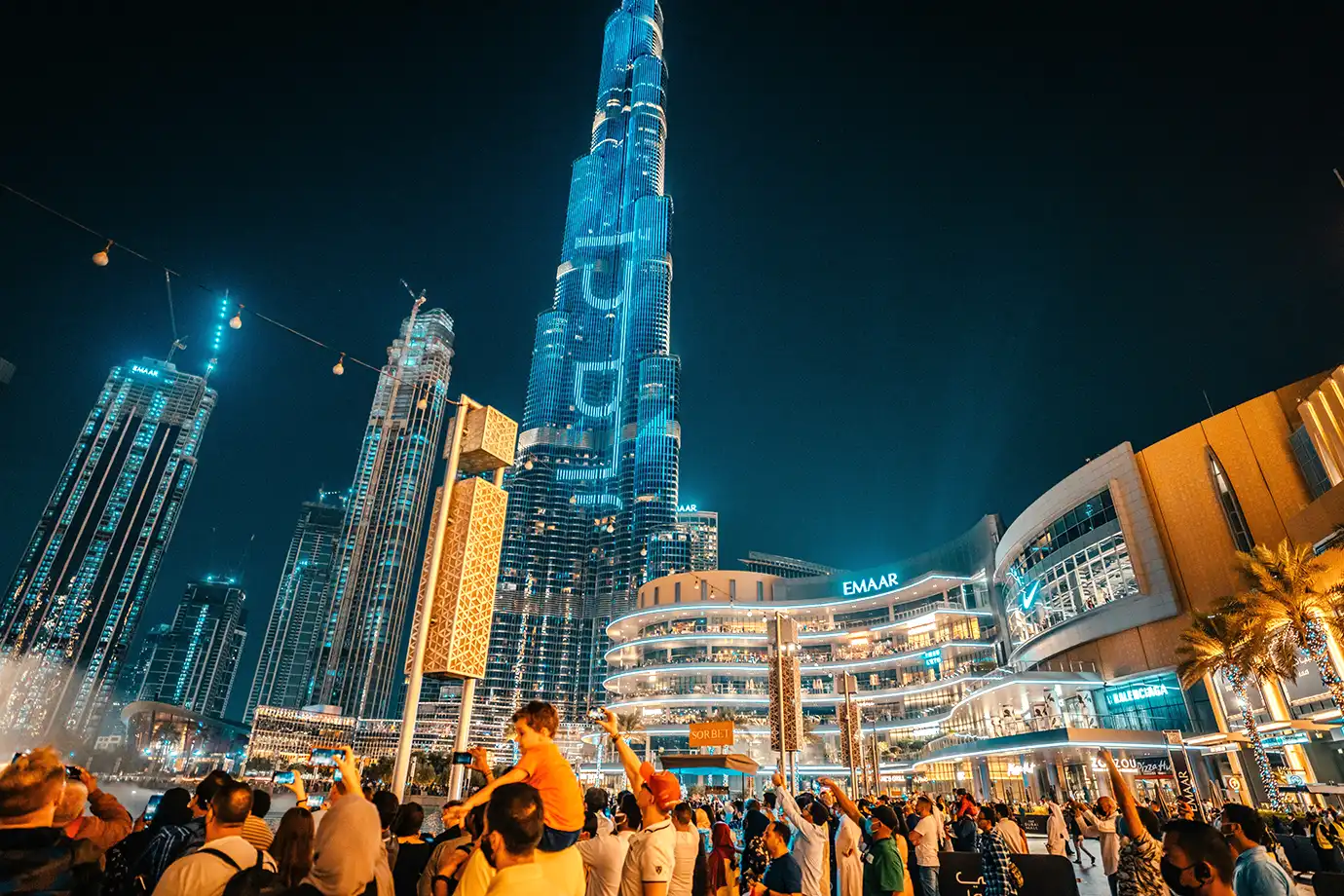 A large crowd gathered at night along the Dubai Mall promenade looking up and taking photos of the illuminated blue Burj Khalifa skyscraper and surrounding urban architecture.