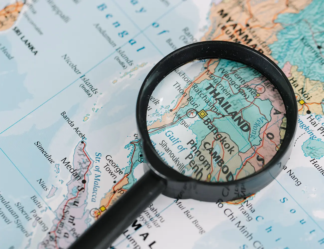 A magnifying glass pointing at Thailand, Cambodia, and Phnom Penh on a printed geographic map featured in a travel guide on Is Cambodia Safe.
