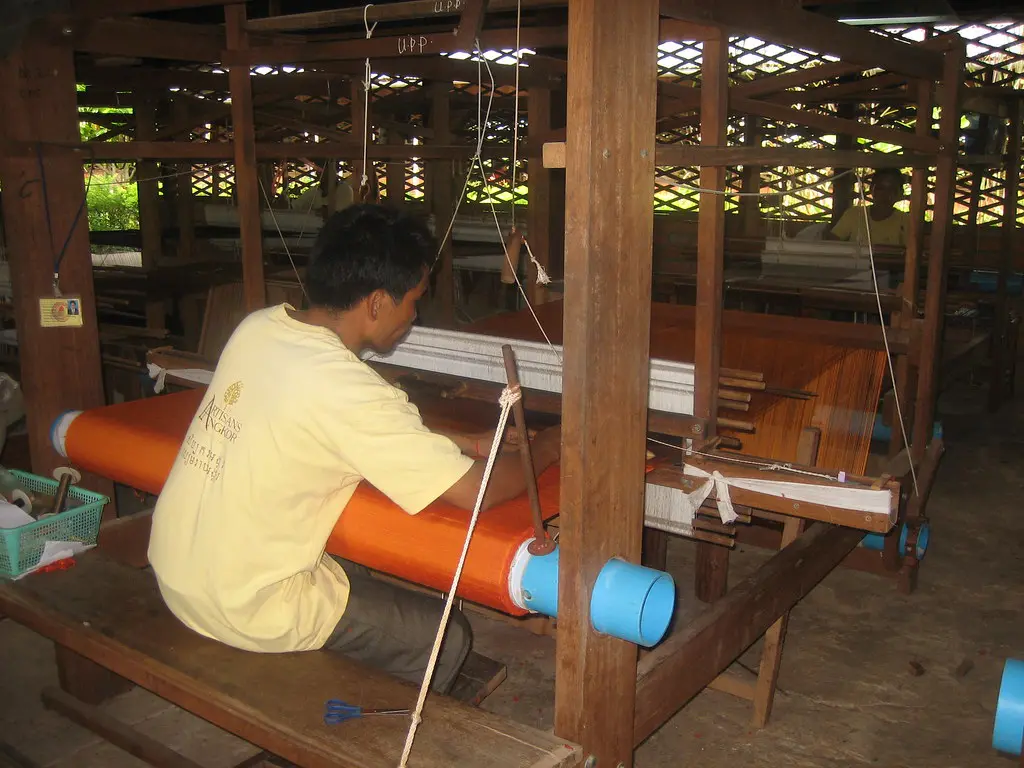 A artisan seated at a traditional wooden handloom weaving bright orange silk fabric featured in a guide on Things to Do in Siem Reap Beyond Angkor Wat.