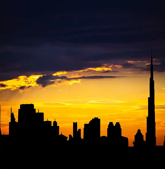 A dramatic black silhouette of the Dubai skyline, anchored by the towering Burj Khalifa, set against a vibrant golden and orange sunset sky with dark clouds overhead.