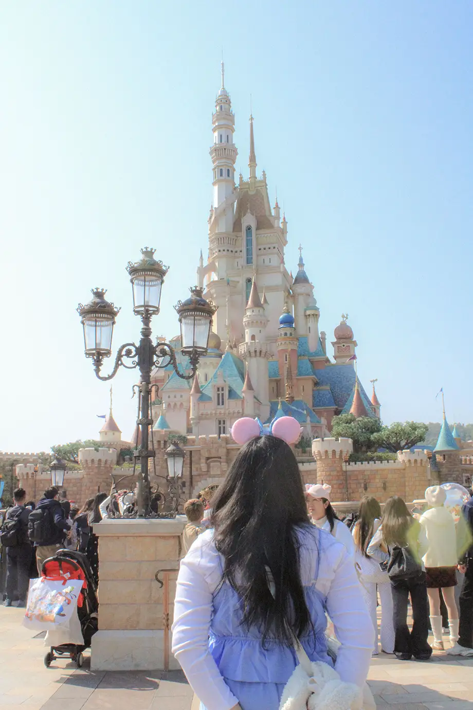 A visitor wearing pink Minnie Mouse ears seen from behind, looking toward the Castle of Magical Dreams with its tall pastel spires and turrets under a clear sky at Hong Kong Disneyland.