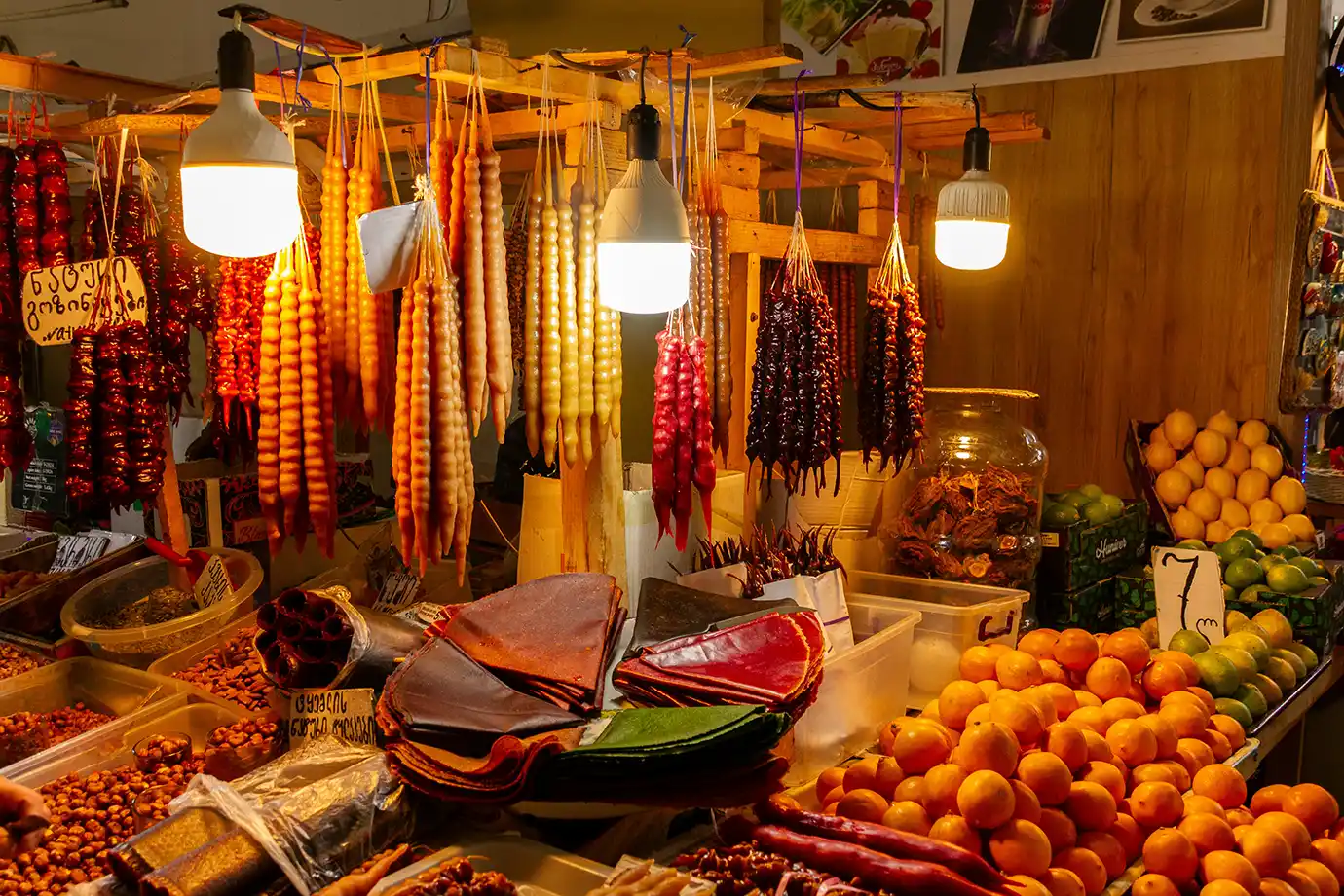 Hanging strands of colorful Churchkhela walnut candies and rolled Tklapi fruit leather at a warm market stall, highlighting sweet snacks for a Georgian Food Guide.