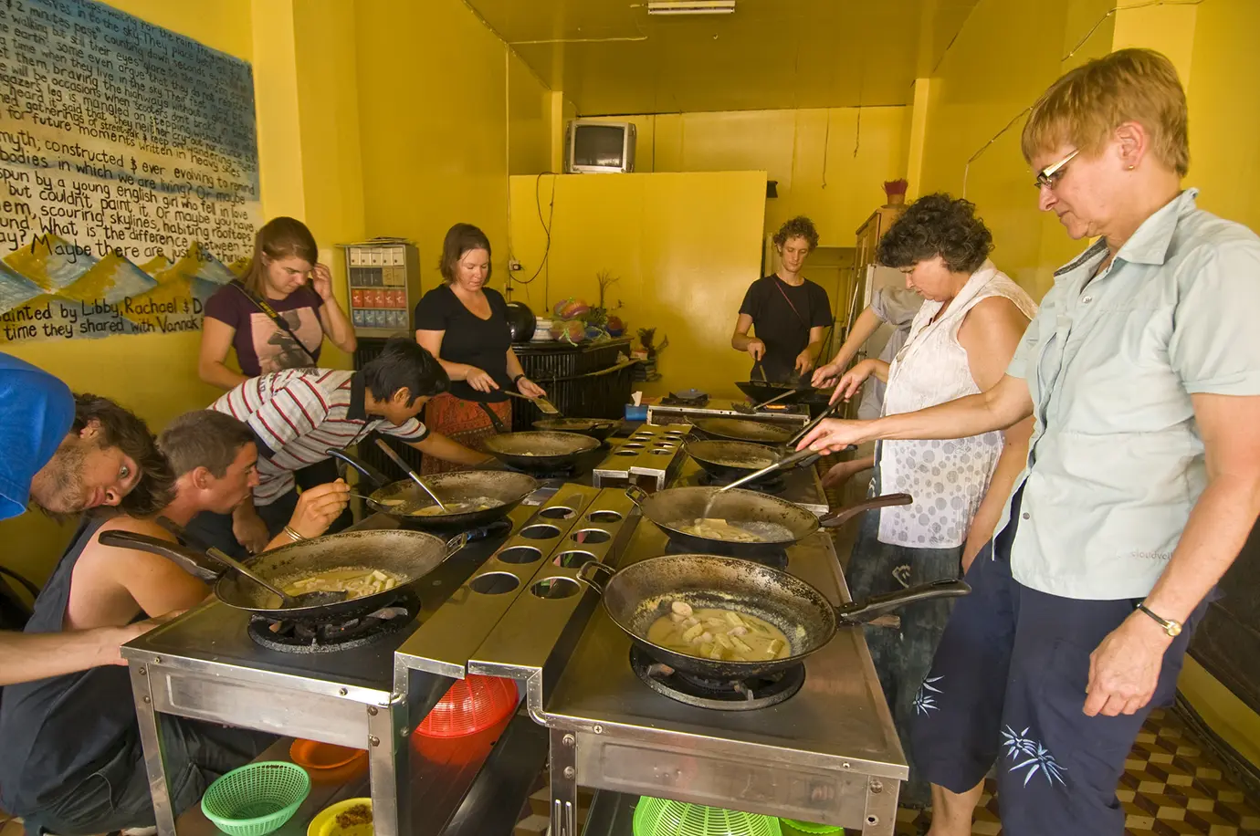 A group of tourists cooking food in individual woks on gas burners during an interactive culinary workshop featured in a guide on Things to Do in Siem Reap Beyond Angkor Wat.