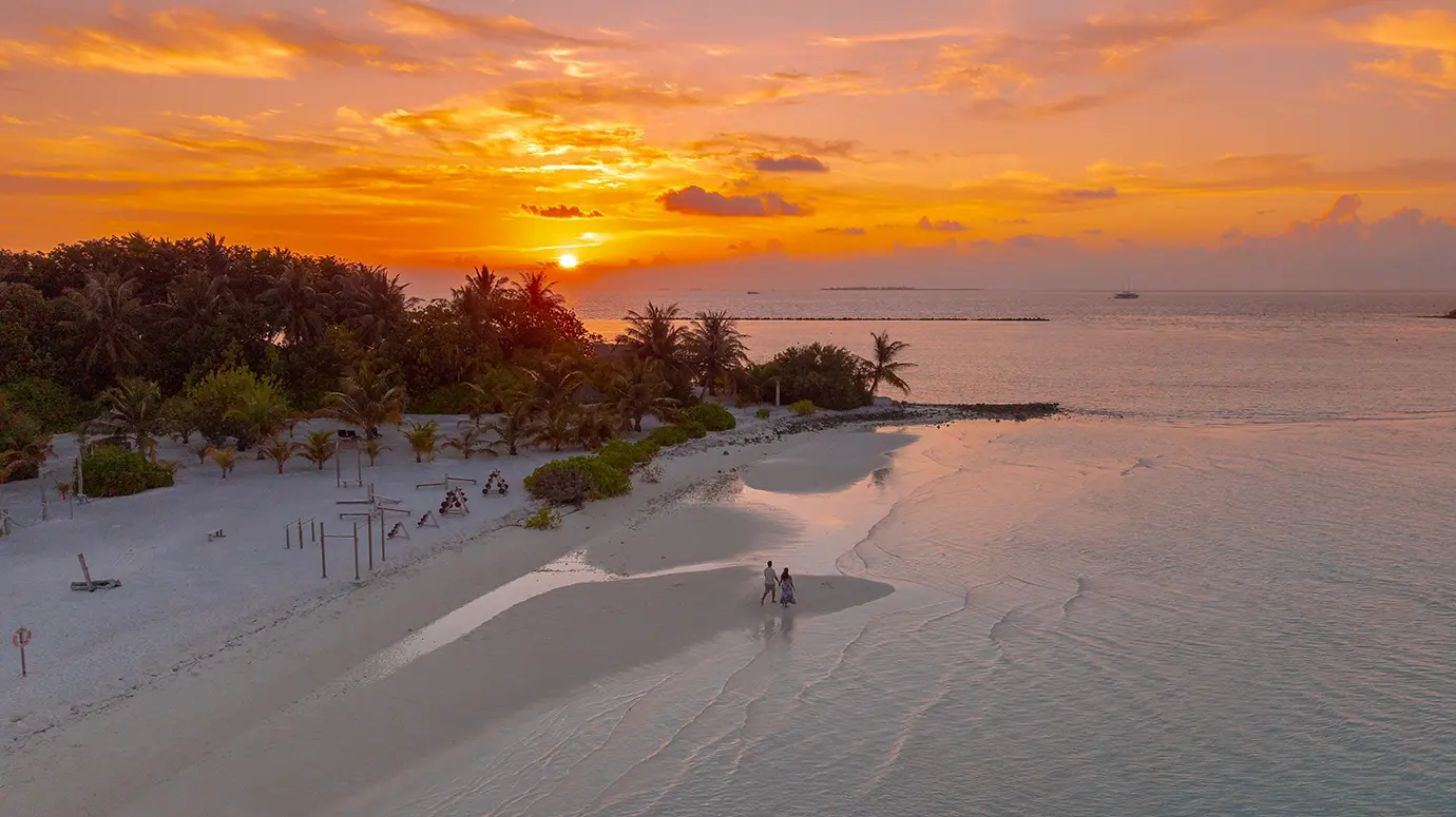 An aerial view of a couple strolling along a white sand sandbank during a vibrant sunset featured in a travel guide on the Best Time to Visit the Maldives.