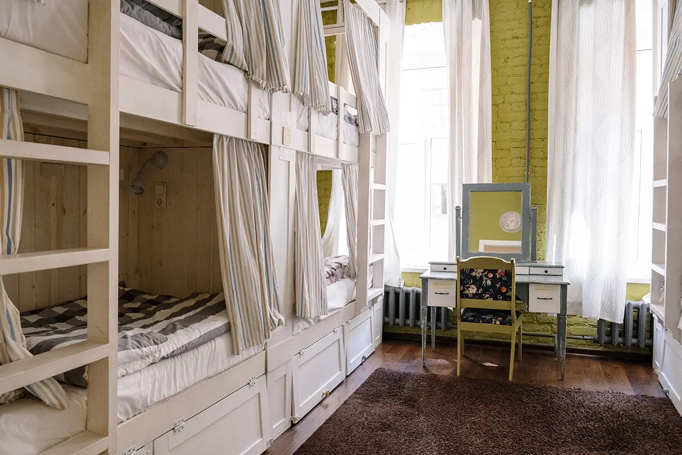 A bright hostel dormitory room featuring wooden bunk beds with privacy curtains, large windows, and a vanity desk, highlighting affordable lodging to minimize Georgia Travel Cost.