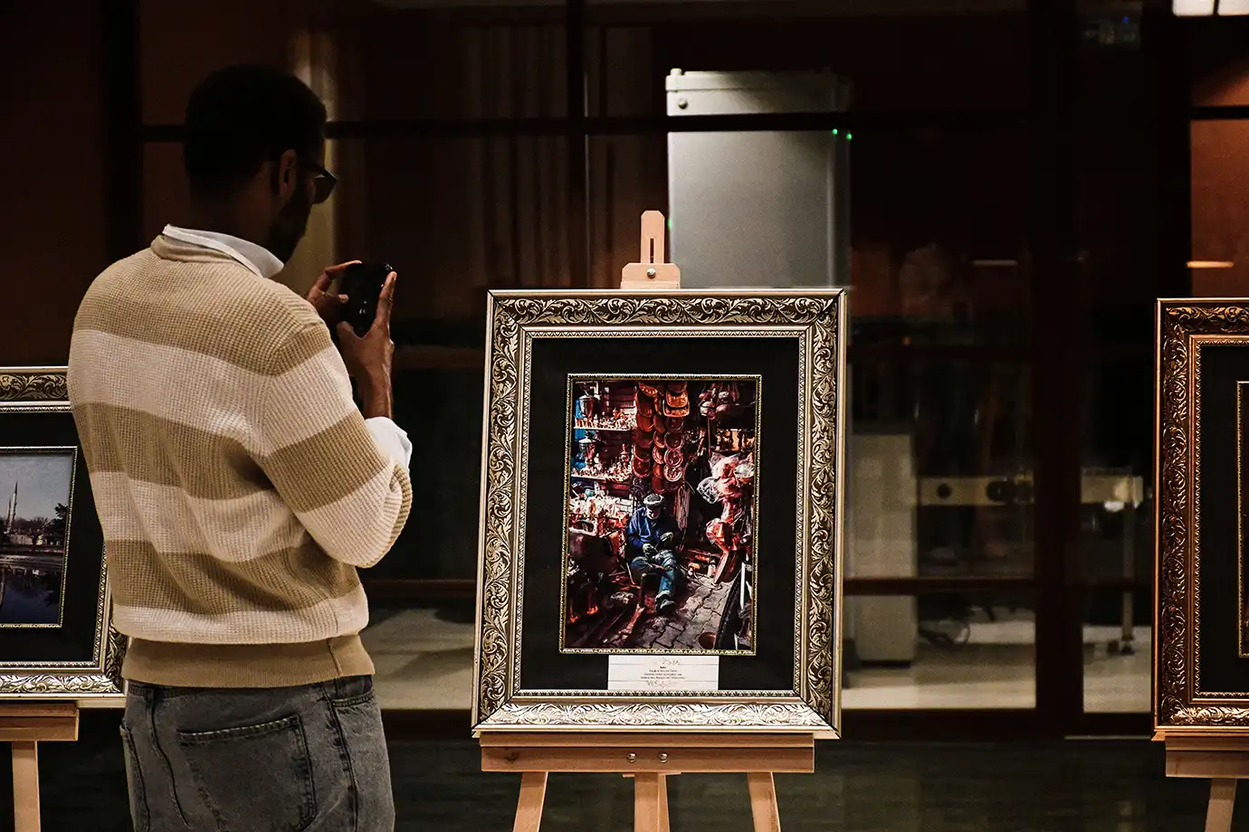 A man in a striped sweater taking a phone picture of framed photography displayed on wooden easels at an indoor art exhibition, featured in Best Time to Visit Dubai.