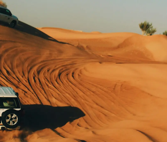 White 4x4 SUVs kicking up sand while driving across steep golden desert dunes during a Dubai Desert Safari.