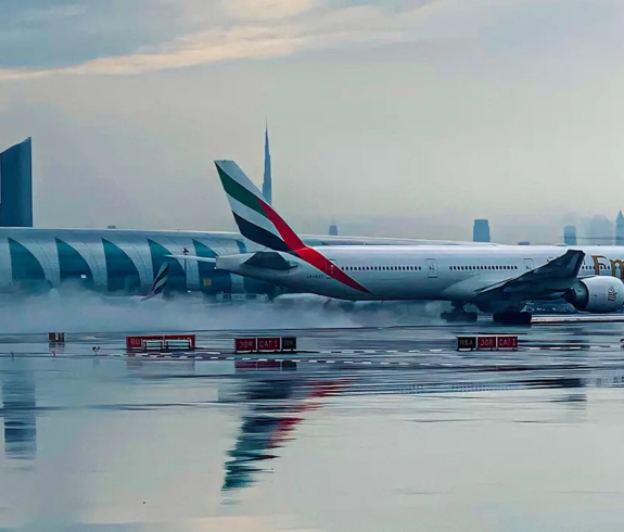 An Emirates Boeing commercial passenger jet taxiing on a wet tarmac at Dubai International Airport with the hazy city skyline in the background, relevant for Dubai Visa Requirements.