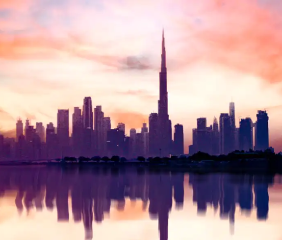 A wide panoramic view of the dark Dubai city skyline and the central Burj Khalifa reflected across calm water under a vibrant pink, orange, and purple sunset sky.