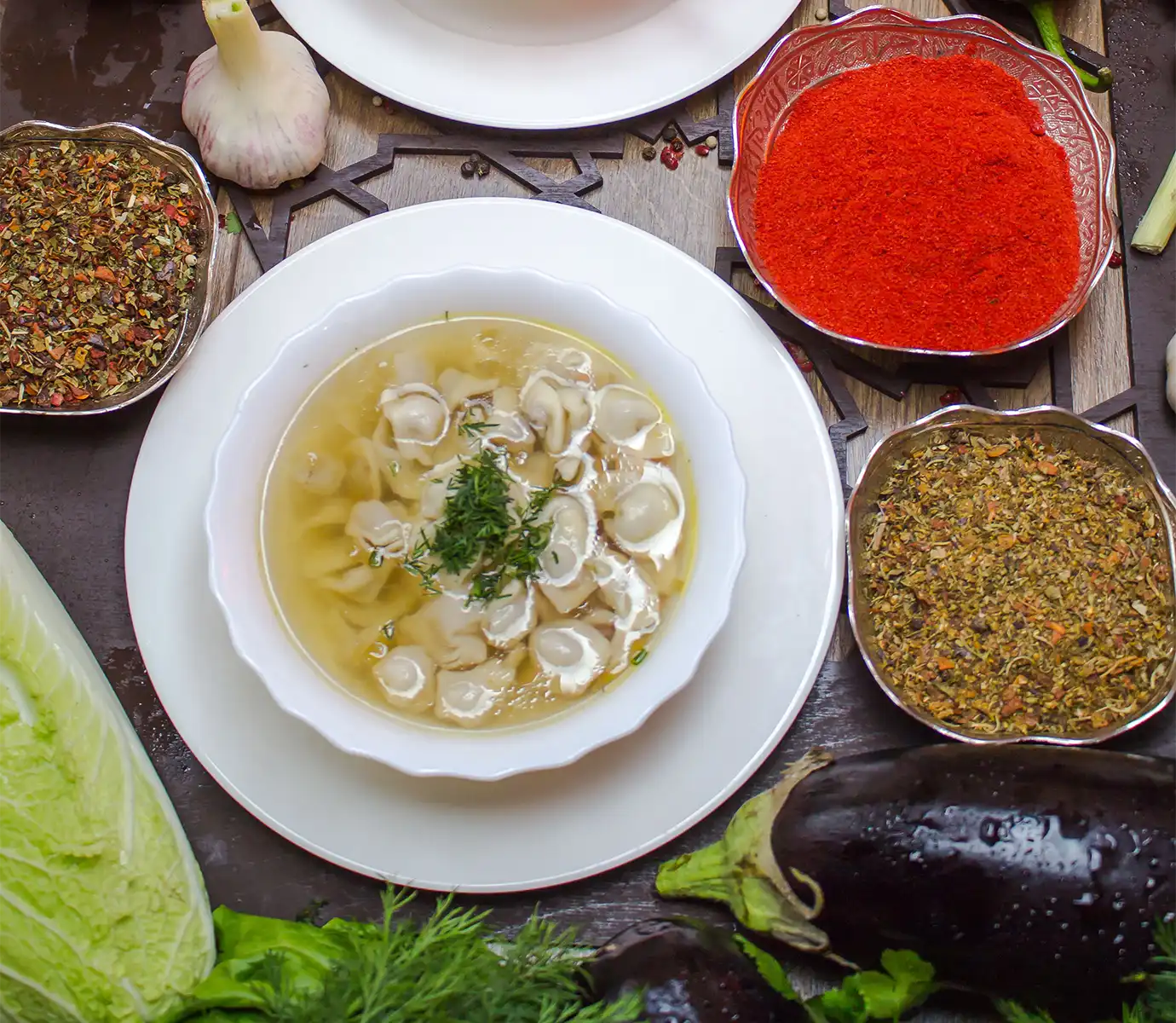 A top-down view of a white bowl containing Azerbaijani dushbara dumpling soup garnished with fresh dill alongside bowls of spices featured in a travel guide on What to Eat in Azerbaijan.