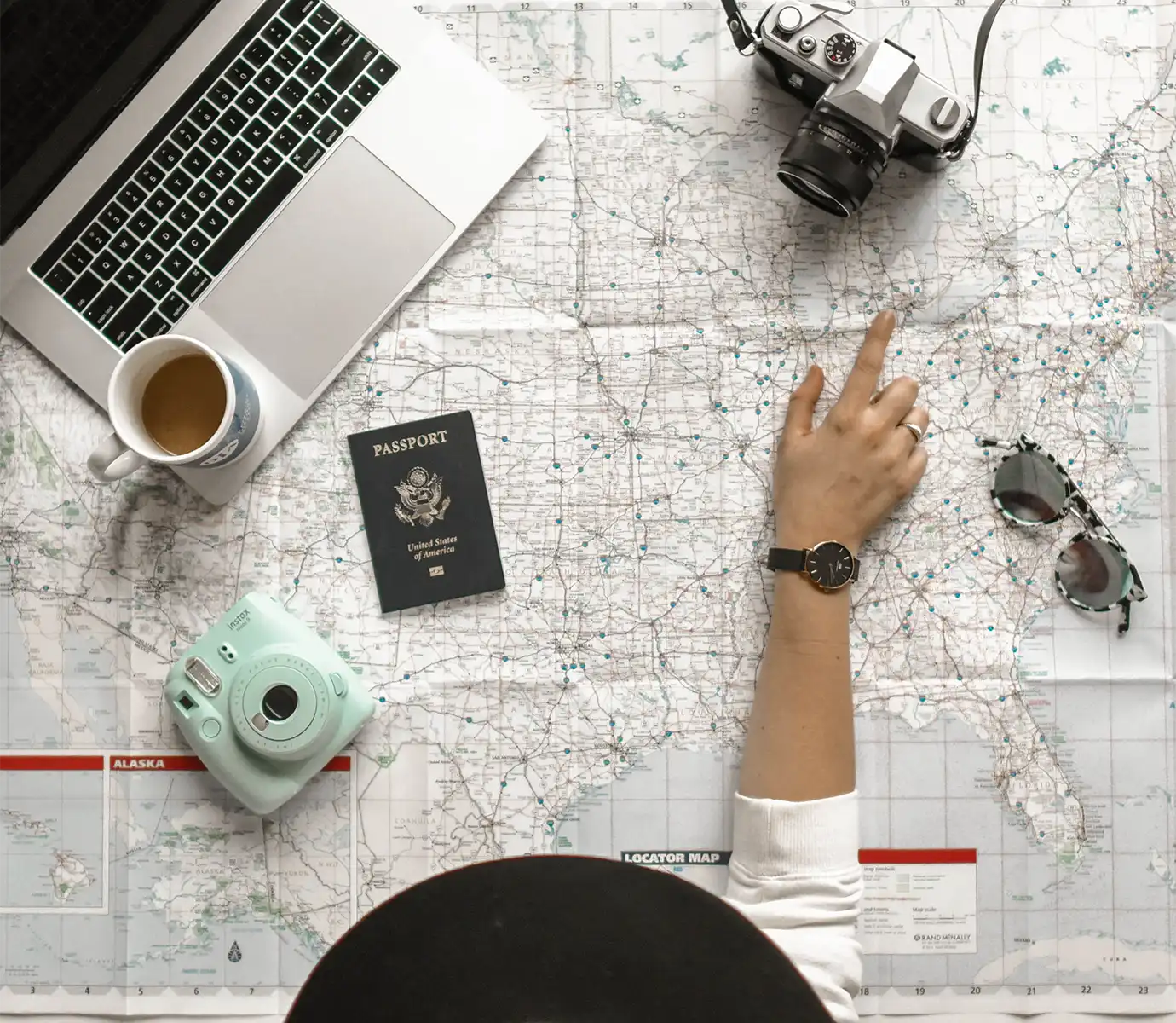 A flat lay overhead view of a US passport, laptop, cameras, coffee cup, and a person's hand pointing at a map featured in a travel guide on Cambodia Visa Requirements.