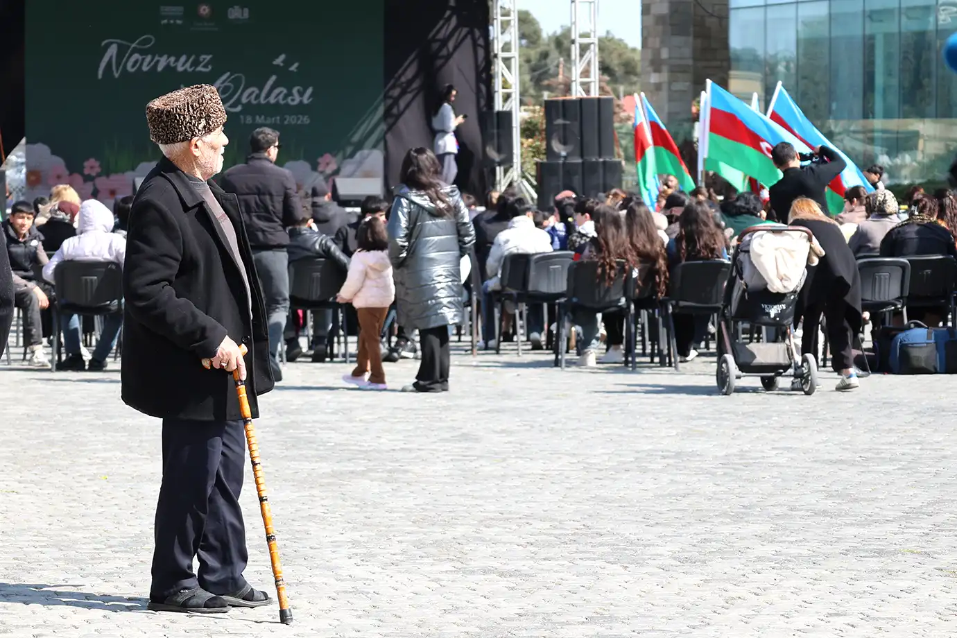 An elderly man in traditional karakul hat leaning on a wooden walking cane while watching a crowd gathered for a Novruz festival with Azerbaijani flags waving in the background.