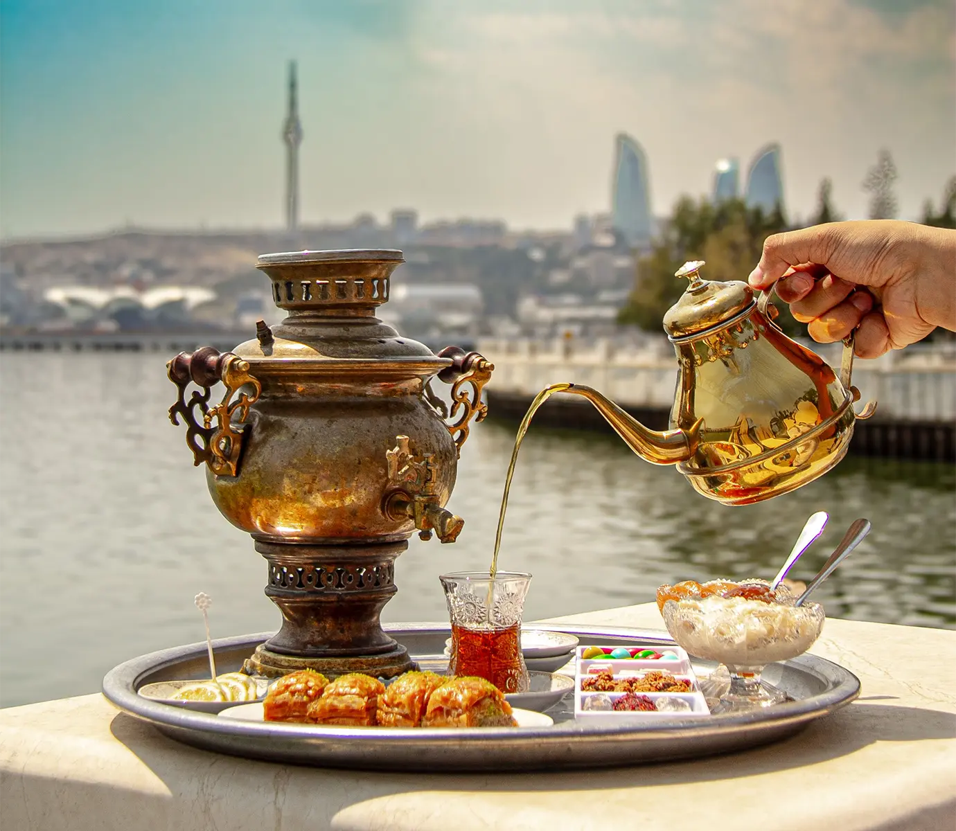 Hot black tea pouring from a brass teapot into an armudu glass next to an antique samovar and sweets along the Baku seafront featured in a travel guide on What to Eat in Azerbaijan.