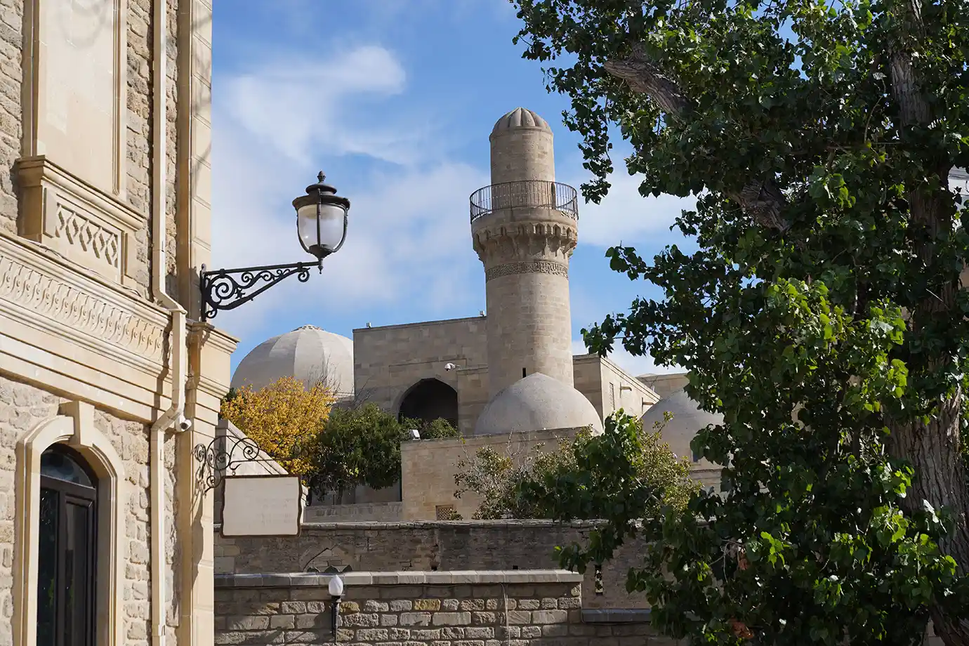 The stone minaret and domed roofs of the Palace of the Shirvanshahs framed by lush green trees and historic architecture in Old City Baku featured in a travel guide on When Is the Best Time to Visit Azerbaijan.
