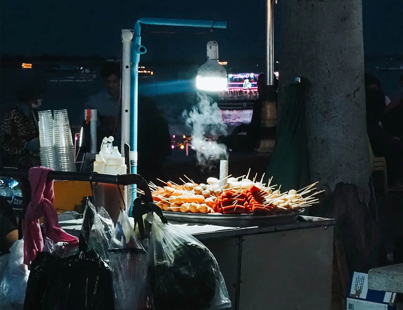 A night street food cart with steam rising from skewers illuminated under a hanging lamp along Phnom Penh's riverfront, featured in a travel guide on Is Cambodia Safe.