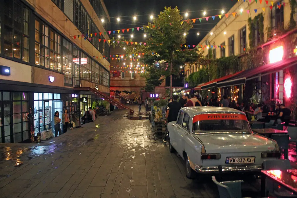 A lively evening scene inside the industrial courtyard of Fabrika Tbilisi, featuring festive string lights, colorful bunting, outdoor cafe seating, and a vintage car, highlighting social Things to Do in Tbilisi.