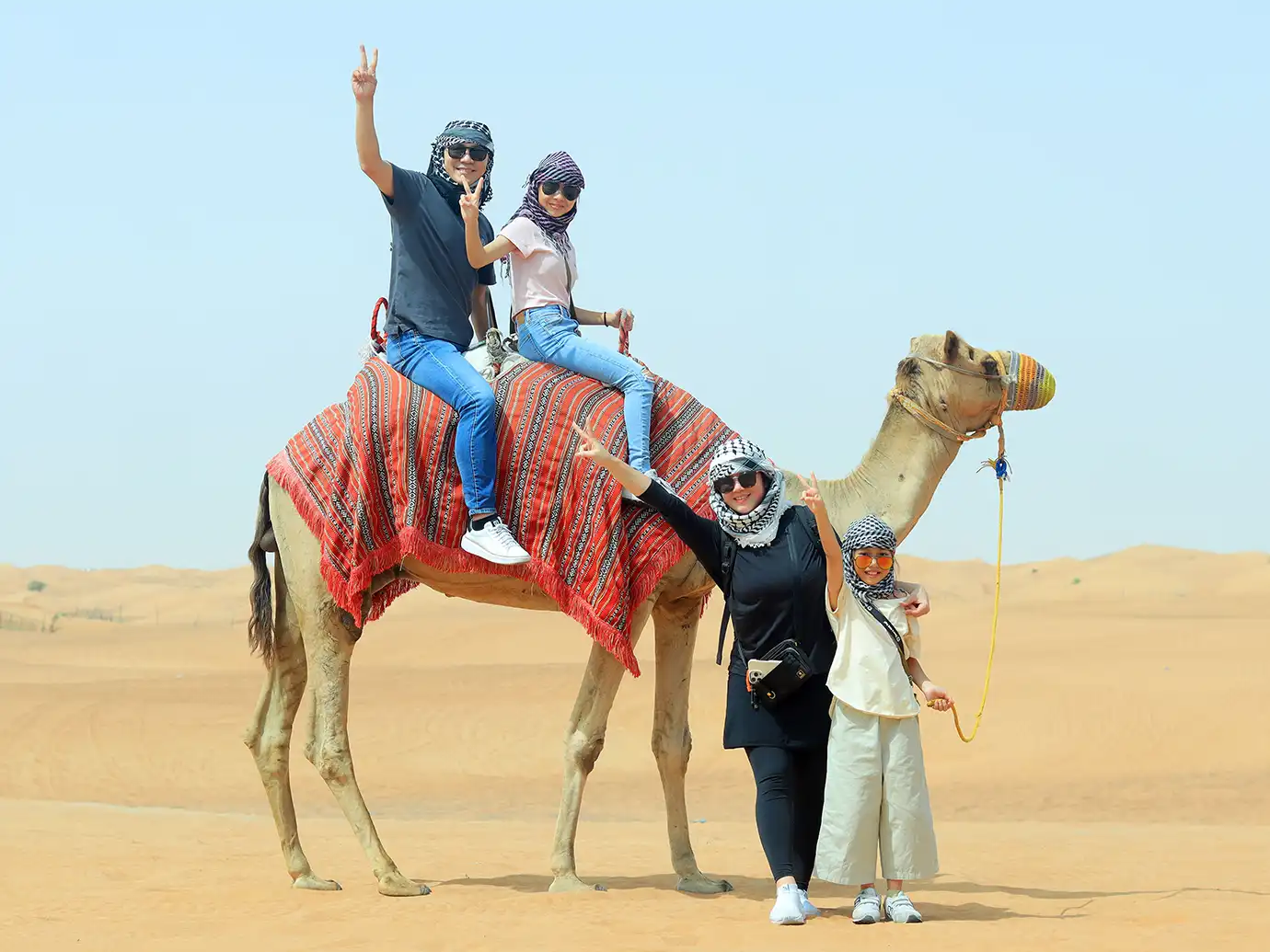 A family of four wearing headscarves and sunglasses posing with a camel on sand dunes, with two family members riding on top and two standing alongside in a desert landscape.