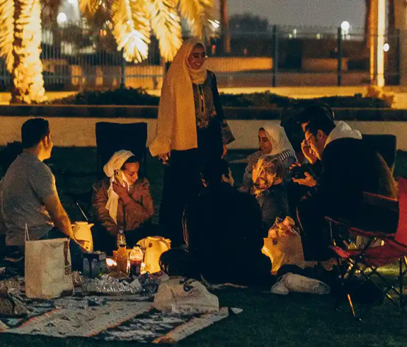 An extended family sitting together on picnic mats and lawn chairs on a green park lawn at night during a trip to Dubai with Kids.