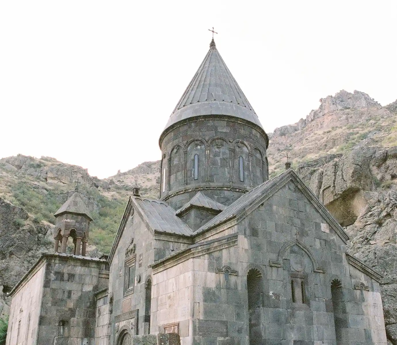 The stone facade and conical cupola of Geghard Monastery built against rugged mountain cliffs featured in a travel guide on 5 Must-See Monasteries in Armenia.