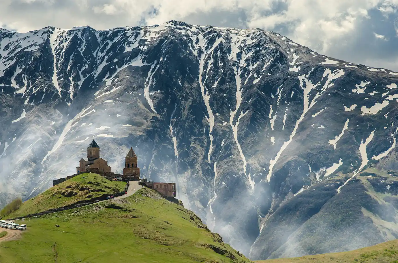 The 14th-century Gergeti Trinity Church standing on a grassy hill beneath massive snow-dusted mountains, showcasing mountain landscapes during the Best Time to Visit Georgia.