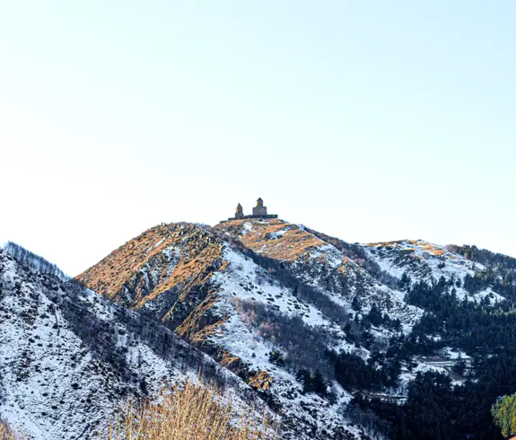 A wide landscape view of the small Gergeti Trinity Church perched atop a hill, framed by snow-dusted mountains and the towering, snow-capped peak of Mount Kazbek under a clear blue sky in Stepantsminda, Georgia.