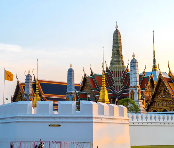 A wide exterior photograph of the white perimeter wall surrounding the Grand Palace complex, with golden spires and tiled roofs of Wat Phra Kaew rising against a sunset sky in Bangkok, Thailand.