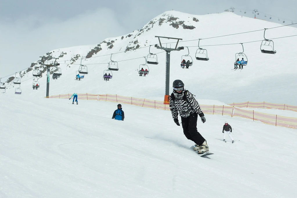 A snowboarder in a patterned jacket riding down a snowy ski slope beneath an active chairlift line at Gudauri Resort, illustrating the Best Time to Visit Georgia.