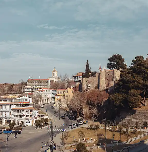 An elevated city view of Old Tbilisi showing traditional buildings with wooden balconies, winding streets, manicured green park areas, and stone fortress walls perched on a rocky cliff under a soft sky.