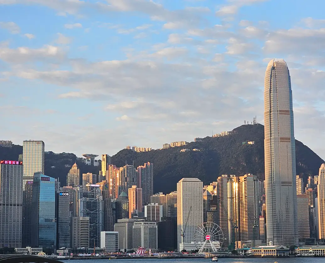 A waterfront view across Victoria Harbour highlighting the Hong Kong Observation Wheel and Two IFC skyscraper illuminated by warm late afternoon sunlight, backed by Victoria Peak under a scattered blue sky.