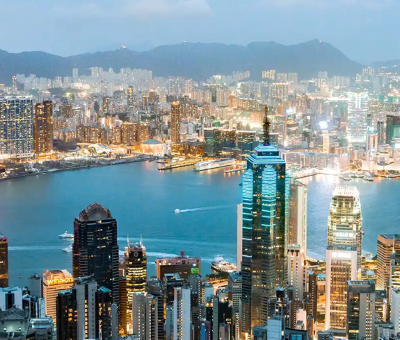 An elevated panoramic view of the glowing Hong Kong skyline at dusk, showing illuminated skyscrapers like Two IFC and ICC surrounding the blue waters of Victoria Harbour.